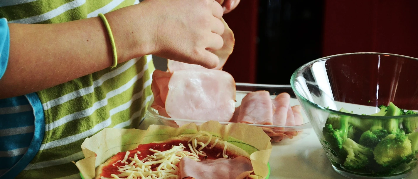 Kochen Das Bild zeigt eine Person, die eine Pizza belegt. Man sieht frischen Brokkoli und geschnittenen Aufschnitt auf dem Tisch.