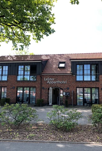 Backsteingebäude des Leister Apparthotels, mit vorgelagerter Terrasse und schattenspendenden Bäumen.