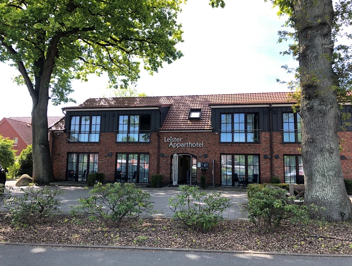 Backsteingebäude des Leister Apparthotels, mit vorgelagerter Terrasse und schattenspendenden Bäumen.