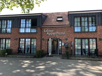 Backstein-Fassade mit zwei Stockwerken, zentralem Eingang und überdachter Terrasse mit Sitzgelegenheiten.