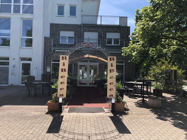 RiBecca-Hotel Der Eingangsbereich des RiBecca-Hotels mit Markise und Pflanzen, umgeben von Bäumen.