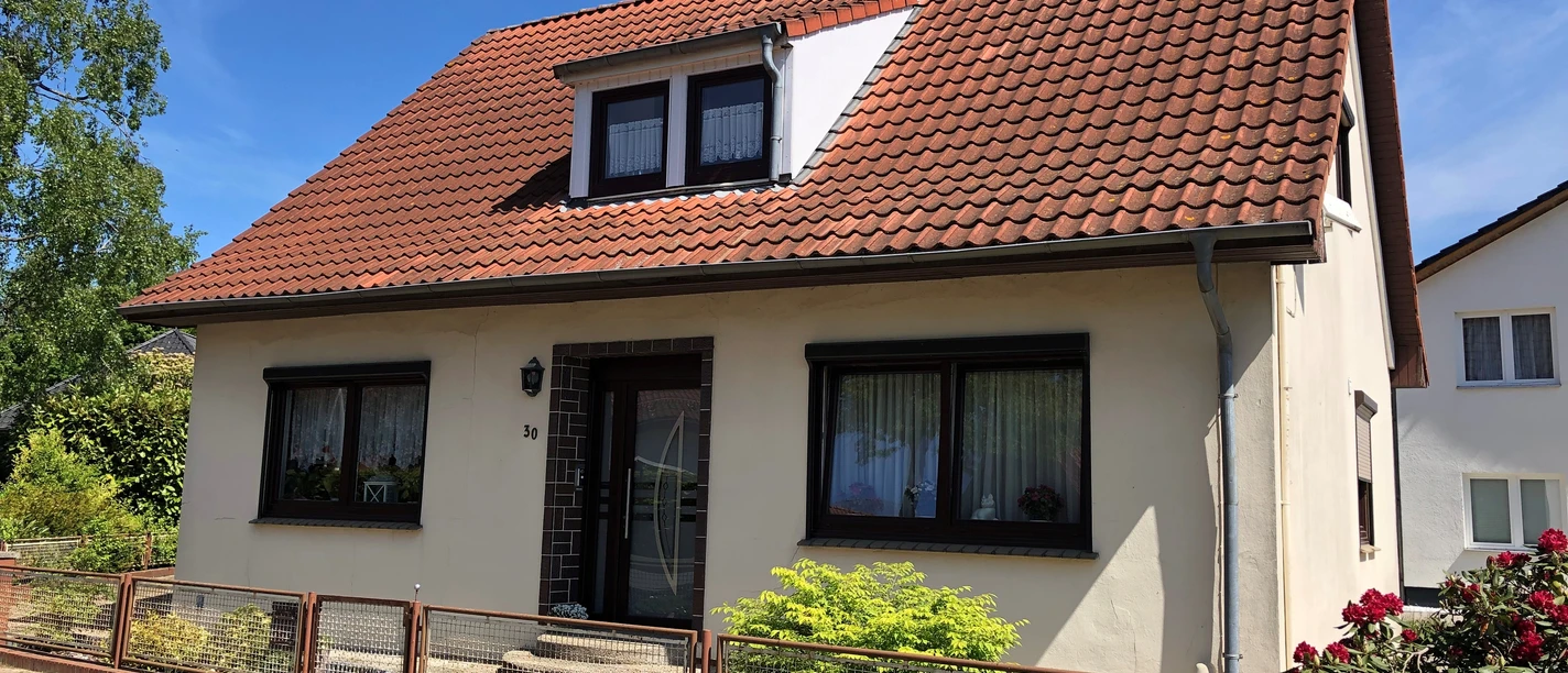 Een vrijstaand huis met een rood zadeldak, omringd door struiken en een speelse voortuin.