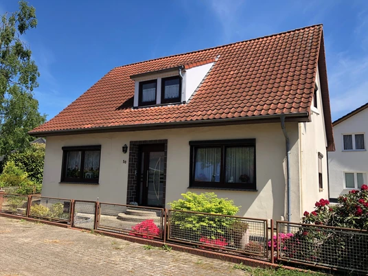 Ruth Fangmann Ein Einfamilienhaus mit rotem Satteldach, umgeben von Büschen und einem verspielten Vorgarten.