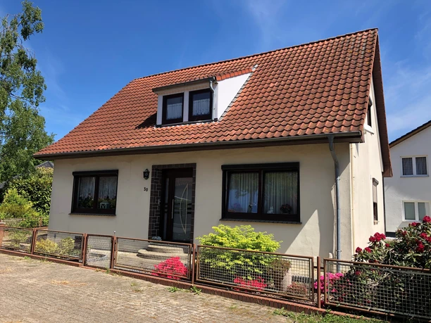 Ruth Fangmann Ein Einfamilienhaus mit rotem Satteldach, umgeben von Büschen und einem verspielten Vorgarten.