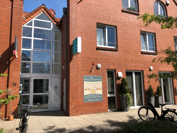 Entrance to a red brick building with a glass façade, bicycles and green plants outside.