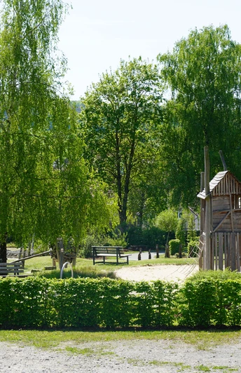 Ein Spielplatz aus Holz, umgeben von üppigen Bäumen und grünen Sträuchern, bietet Platz zum Entdecken.