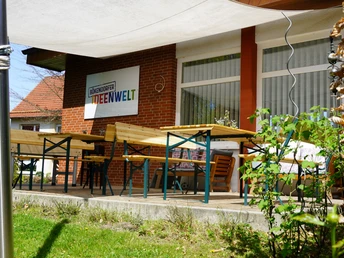 Ideenwelt 2.JPG Außenansicht eines Cafés mit Terrasse, Tischen und Stühlen vor einem Ziegelgebäude unter Sonnensegel.