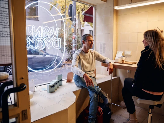VAN DYCK Espressobar Ein Mann und eine Frau sitzen entspannt in der VAN DYCK Espressobar, hinter ihnen ist ein großes, weiß beleuchtetes Fenster mit dem Logo der Bar zu sehen. Die Atmosphäre wirkt warm und einladend.A man and a woman are sitting relaxed in the VAN DYCK espresso bar, behind them a large, brightly lit window displays the café's logo. The atmosphere appears warm and inviting.