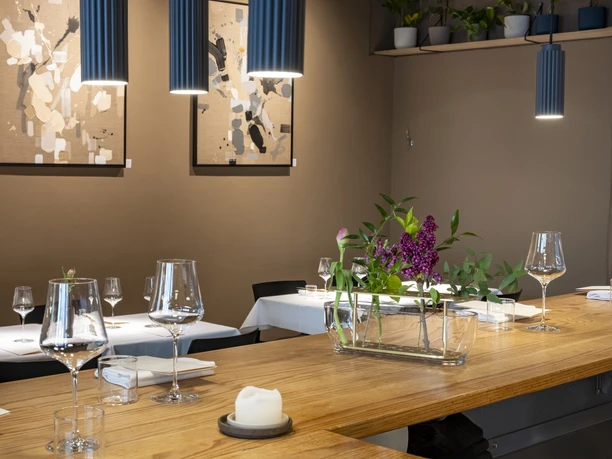 Pottkind The image shows an elegant, modern restaurant interior featuring tasteful table decor, artistic wall prints, and harmonious lighting. In the foreground, a decorative flower arrangement in a glass vase is placed on a wooden bar counter.