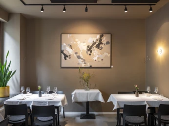 Pottkind In einem stilvoll eingerichteten Restaurant stehen zwei gedeckte Tische vor einer abstrakten Kunstwand.In a stylishly decorated restaurant, two set tables stand in front of a wall displaying abstract art.