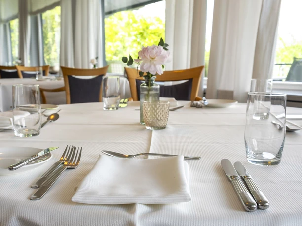 Zur Tant An elegantly set table in a bright, light-filled restaurant. White tablecloth with cutlery, glasses, and a floral vase arrangement.
