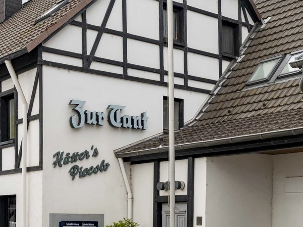 Zur Tant A half-timbered house with the inscription "Zur Tant". The building is white with dark timber beams, a chimney, and has an entrance with a door and shop windows.
