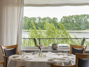 Zur Tant Ein stilvoll eingedeckter Tisch vor einem großen Fenster mit Blick auf einen Fluss und grüne Bäume.A stylishly set table in front of a large window with a view of a river and green trees.