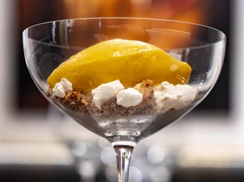 OTTO für dich Ein elegantes Dessert in einem hohen, klaren Glas. Die Hauptkomponente ist ein leuchtend gelbes Sorbet, das auf einer Schicht aus Krümeln und kleinen, weißen Baiser-Stücken liegt. Der Hintergrund ist unscharf, was den Fokus auf das ansprechend präsentierte Dessert verstärkt.An elegant dessert served in a tall, clear glass. The main component is a vibrant yellow sorbet, resting on a layer of crumbles and small pieces of white meringue. The background is blurred, enhancing the focus on the attractively presented dessert.
