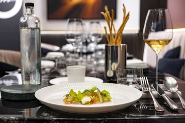 OTTO für dich Ein elegant gedeckter Tisch, auf dem ein kunstvoll angerichtetes Gericht mit Kräutergarnitur serviert wird, daneben ein Glas Weißwein und stilvolles Besteck.