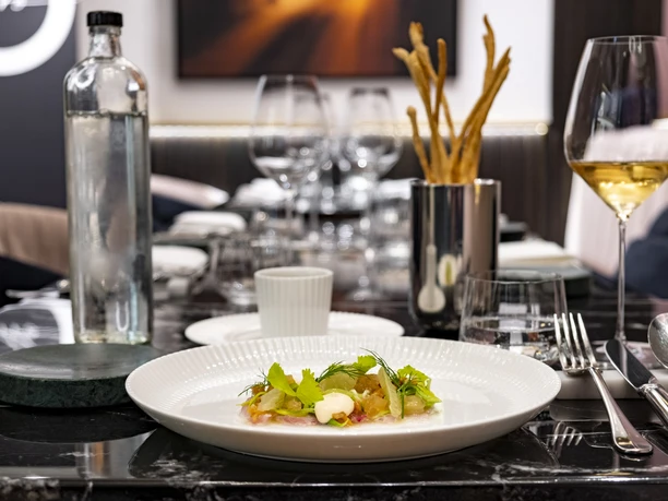 OTTO für dich An elegantly set table, serving an artfully plated dish garnished with herbs, accompanied by a glass of white wine and stylish cutlery.