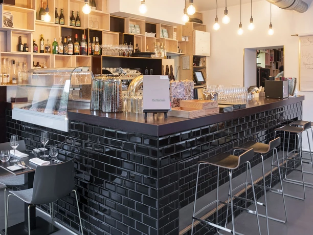 maiBeck Einladende Theke in einem modernen Restaurant. Die Theke ist mit glänzenden schwarzen Fliesen verkleidet und umrahmt eine Auswahl an Weinen, Gläsern und dekorativen Elementen. An der Theke befinden sich stilvolle Barhocker, während im Vordergrund ein gedeckter Tisch für zwei Gäste bereitsteht. Der Raum ist durch sanfte Beleuchtung und hängende Glühbirnen an der Decke atmosphärisch gestaltet.