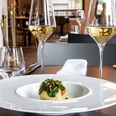 maiBeck Elegantes Restaurantinterieur mit stilvoll gedecktem Tisch, fokussiert auf ein exquisites Gericht mit frischen, grünen Zutaten, flankiert von zwei Gläsern Weißwein. Elegant restaurant interior featuring a stylishly set table, centered around an exquisite dish with fresh, green ingredients, accompanied by two glasses of white wine.