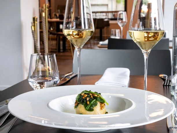 maiBeck Elegantes Restaurantinterieur mit stilvoll gedecktem Tisch, fokussiert auf ein exquisites Gericht mit frischen, grünen Zutaten, flankiert von zwei Gläsern Weißwein.