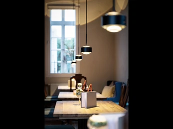 002_Alte-Remise_Urheber_Top-Light.jpg Ein stilvoll eingerichtetes Café mit Holztischen und stimmungsvoller Beleuchtung neben einem Fenster.