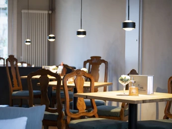 003_Alte-Remise_Urheber_Top-Light.jpg Holzstühle und -tische in einem stilvoll beleuchteten Raum, vermittelt durch wandhängende Lampen.