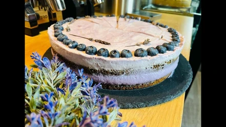 Heidelbeer-Lavendel-Torte auf schwarzer Platte, dekoriert mit Lavendel und Blaubeeren, neben Espressomaschine.