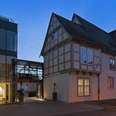 Abendliche Außenansicht eines modernen Gebäudetrakts neben einem historischen Fachwerkhaus.