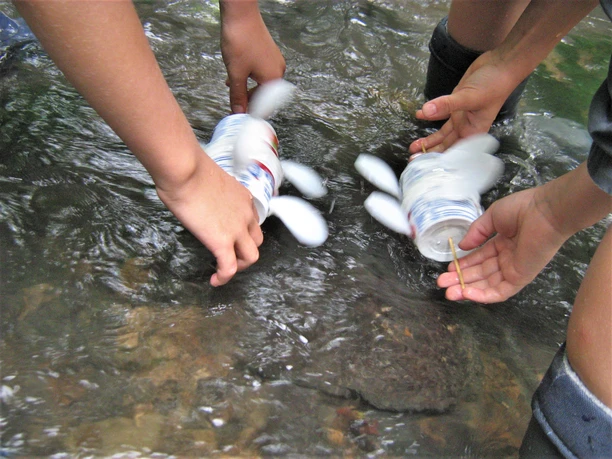 Wasserbewegt Kleine Mühlen Kinder mit Gummistiefeln lassen selbstgebastelte Boote aus Flaschen auf einem flachen Bach schwimmen.