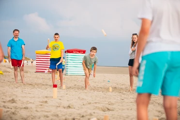 Some children and adults play an outdoor throwing game on the sandy beach under a blue sky.