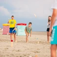 Some children and adults play an outdoor throwing game on the sandy beach under a blue sky.