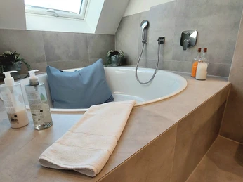 Helles Bad mit Whirlpoolwanne, grauen Fliesen, Fenster, Deko-Pflanzen, Handtuch und Pflegeprodukten.