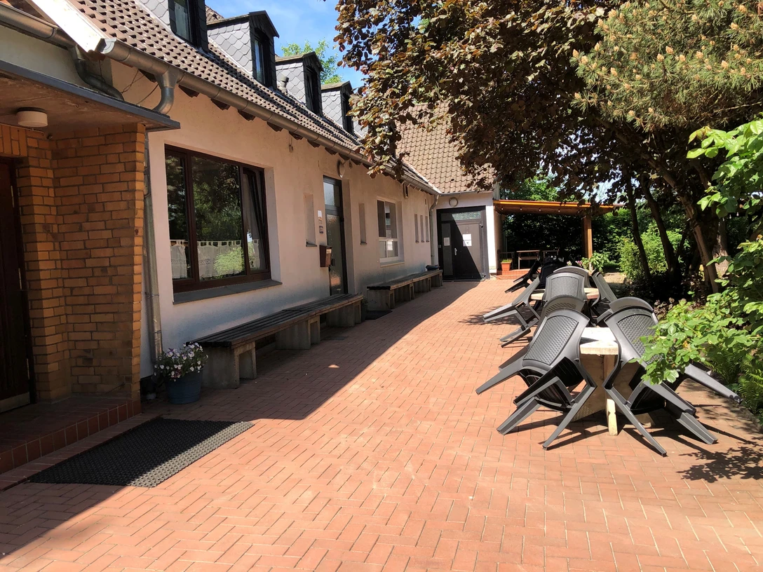 Außenansicht Das Bild zeigt einen gepflasterten Außenbereich mit Bäumen, Bänken und mehreren schwarzen Stühlen.The picture shows a paved outdoor area with trees, benches and several black chairs.Billedet viser et asfalteret udendørs område med træer, bænke og flere sorte stole.Op de foto zie je een geplaveide buitenruimte met bomen, banken en een aantal zwarte stoelen.