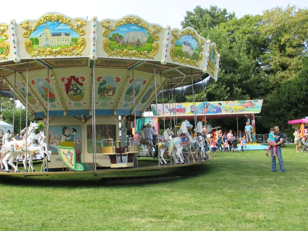 zwiwo-karussell Ein nostalgisches Karussell mit kunstvoll bemalten Dächern und weißen Pferden dreht sich auf einer grünen Wiese. Im Hintergrund sind Besucher und bunte Buden zu sehen.