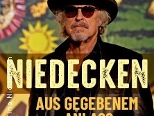 Sänger Wolfgang Niedecken in Sonnenbrille und Hut vor farbenfrohem Hintergrund mit Sonnendesign.