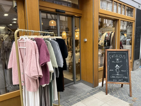 No.Five Eingang Geschäftseingang, davor steht ein Kleiderständer mit flauschigen Oberteilen und ein Schild auf dem steht "Fashion-Alarm Deine neuen Lieblingsstücke sind da!"