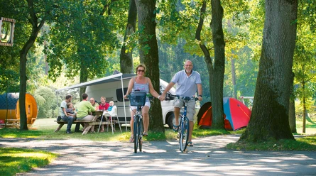 Ein fröhliches Paar radelt auf einem bewaldeten Campingplatz an vorbei sitzenden Menschen.