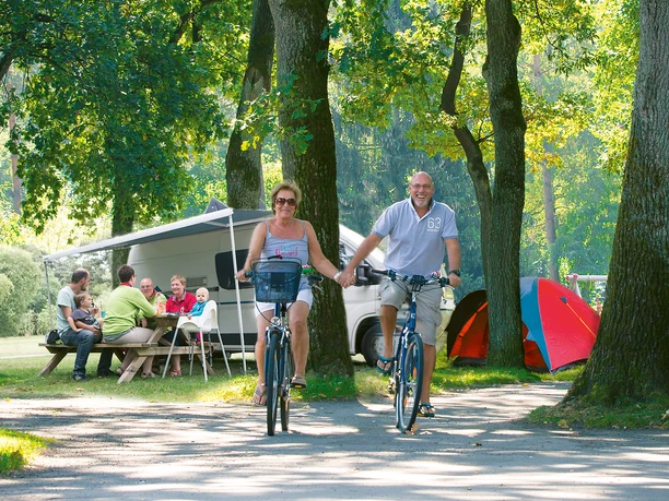 Ein fröhliches Paar radelt auf einem bewaldeten Campingplatz an vorbei sitzenden Menschen.