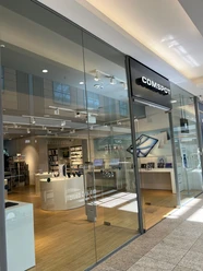 COMSPOT City Galerie Wolfsburg Die Fassade eines modernen Elektronikgeschäfts mit Glasfront, benannt "COMSPOT", im Einkaufszentrum. Innen sieht man helle Holzfußböden und Ausstellungsflächen.