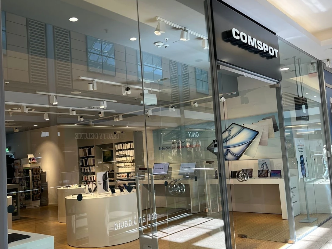 COMSPOT City Galerie Wolfsburg Die Fassade eines modernen Elektronikgeschäfts mit Glasfront, benannt "COMSPOT", im Einkaufszentrum. Innen sieht man helle Holzfußböden und Ausstellungsflächen.