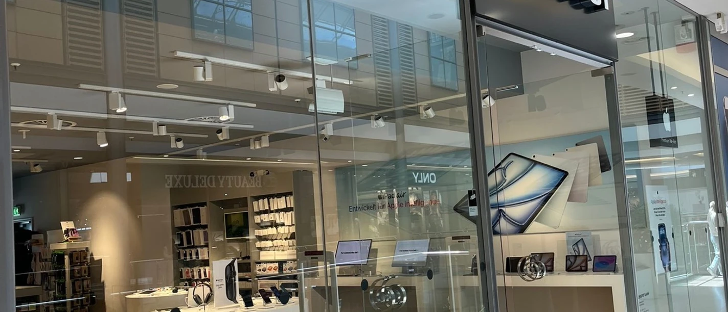 COMSPOT City Galerie Wolfsburg Die Fassade eines modernen Elektronikgeschäfts mit Glasfront, benannt "COMSPOT", im Einkaufszentrum. Innen sieht man helle Holzfußböden und Ausstellungsflächen.