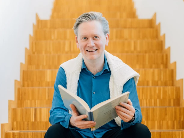 Jonathan Bloxham sitzt lächelnd auf einer Holztreppe, hält ein aufgeschlagenes Buch in den Händen.