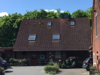Een rood bakstenen huis met een groot schuin dak en kleine dakramen tegen een weelderig groene bosrijke achtergrond.