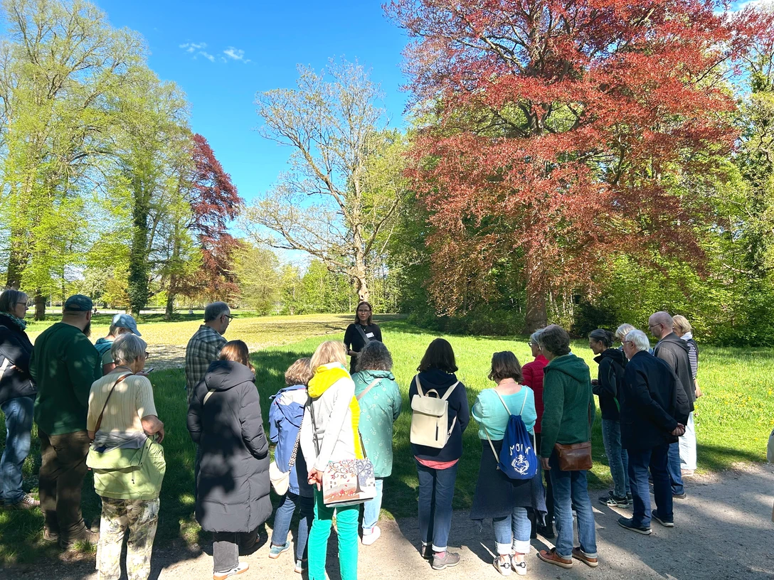 Kräuterwanderung im Rasteder Schlosspark Gruppe lauscht einer Kräuterführerin im sonnigen Schlosspark Rastede zwischen grünen und roten Bäumen.