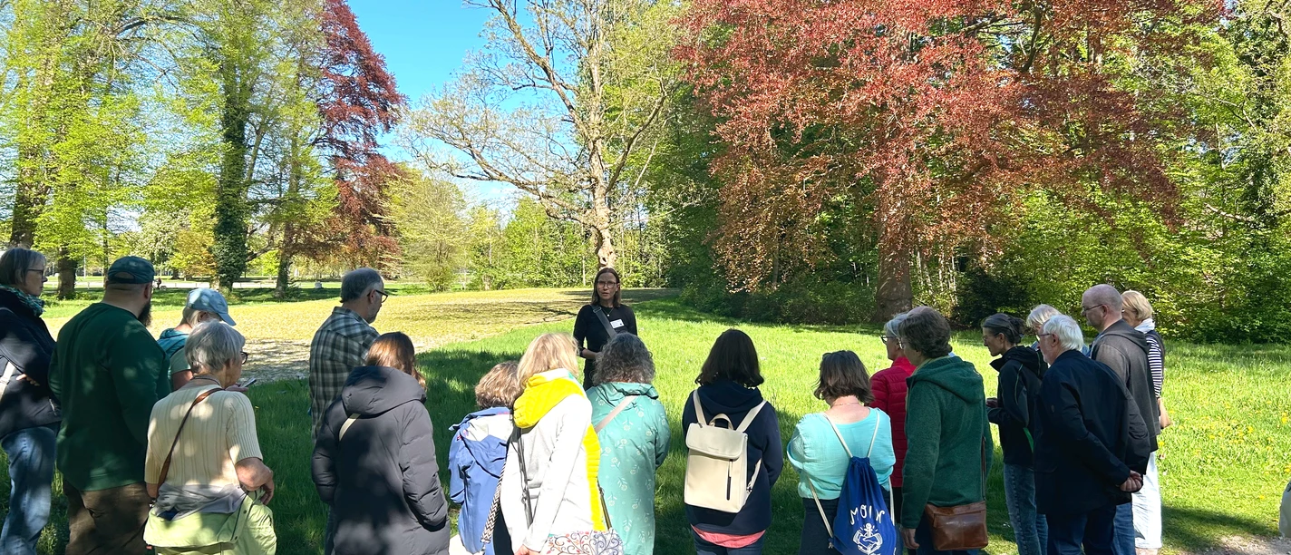 Kräuterwanderung im Rasteder Schlosspark Gruppe lauscht einer Kräuterführerin im sonnigen Schlosspark Rastede zwischen grünen und roten Bäumen.
