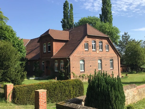 A red brick building with a pitched roof stands in front of a hedge, surrounded by green trees and lawn.