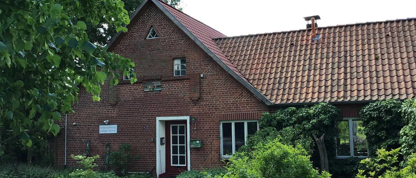 FeWo Strump Rotes Backsteinhaus mit rotem Ziegeldach und begrüntem Garten, umgeben von üppiger Vegetation.