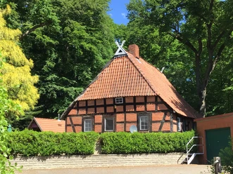 A traditional half-timbered house with a red tiled roof surrounded by lush greenery in a rural setting.