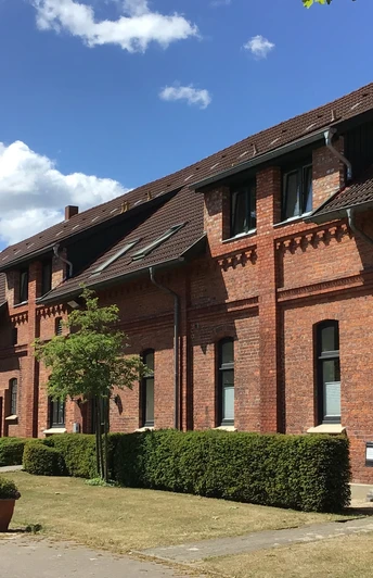 Gästehaus Schlossgarten Backsteingebäude mit markanten Giebeln und gepflegtem Vorgarten bei blauem Himmel und Sonnenschein.