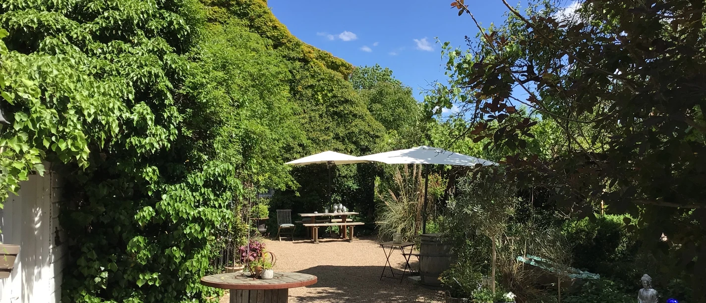 Een groen landhuis met weelderige klimplanten, parasols en een pad in fijne kiezel in de tuin.