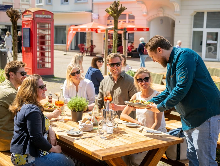 Herford-2025-155-©Teutoburger Wald - Pro Herford GmbH - D. Ketz.jpg Eine Gruppe genießt ein Essen im Freien an einem Holztisch unter Sonnenschirmen in einer Stadt.